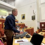 Jussi Kananen seisoo ja puhuu valtuustossa. Tumman sininen paita. Vaalea tukka. Ruskeat housut. Mikrofoni edessä ja tietokone. Taustalla näkyy nouseva katsomo.