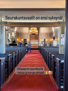 kuva on viitasaaren kirkosta sisältä. siinä näkyy keskellä punainen matto ja reunoilla siniset penkit. Perällä on alttaritaulu ja siellä suunnalla kolme ihmistä keskustelee