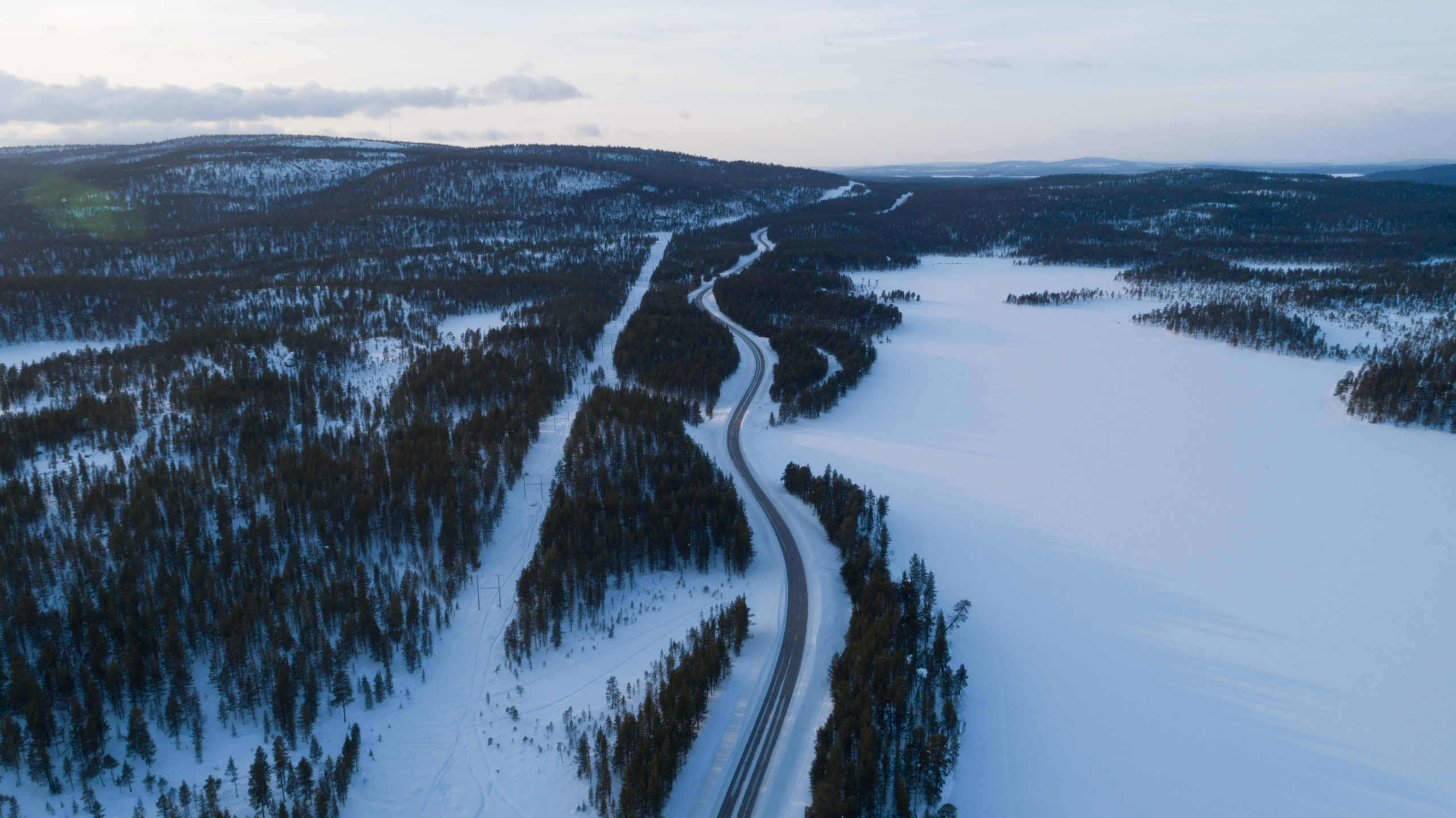 Korkealta kuvattu luminen valtatiemaisema. oikeallä jäinen ja luminne järvi. Vasemmalla metsää ja kaksi tunturia.